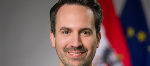 Ein Porträt von Bildungsminister Christoph Wiederkehr mit freundlichem Blick - im Hintergrund sind verschwommen die Flaggen von Österreich und der Europäischen Union zu sehen.
