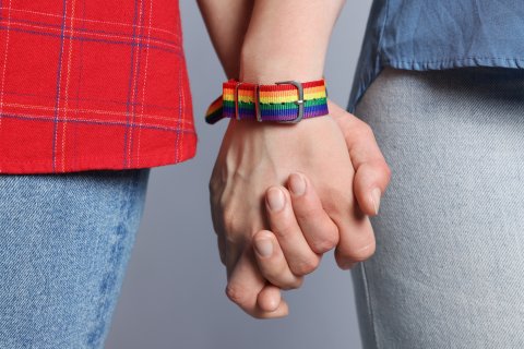 Zwei Hände mit einem Regenbogenarmband halten einander fest.