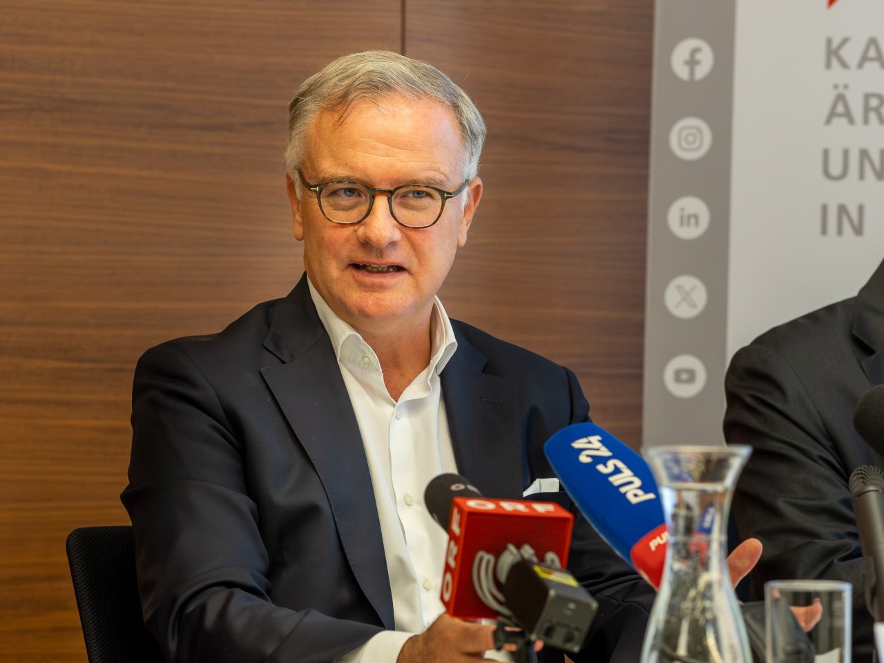 Ein Mann spricht im Rahmen einer Pressekonferenz, vor ihm stehen Mikrophone von Radio- und Fernsehanstalten.