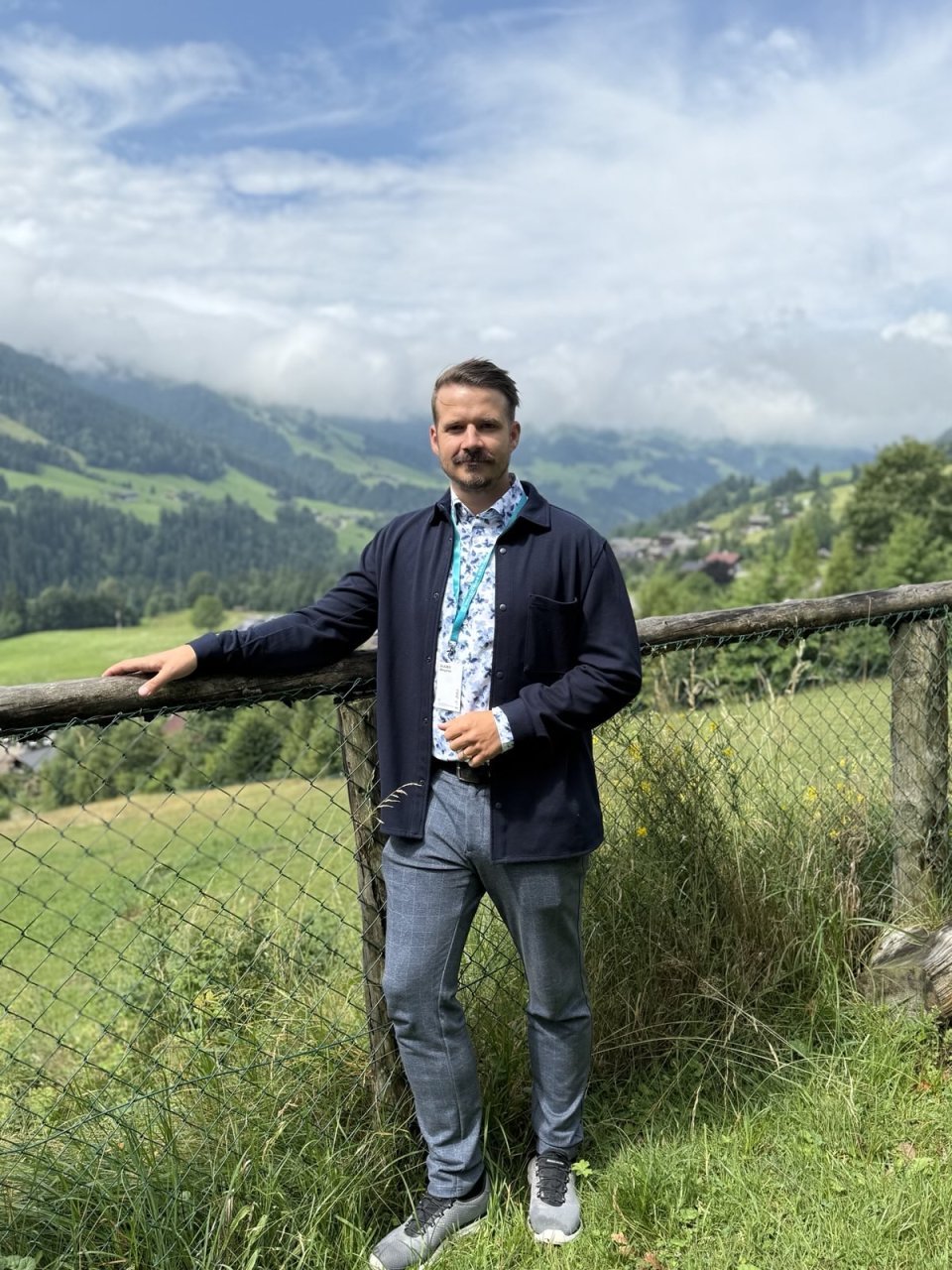 Benjamin Glaser, Referent in der Kammer für Ärztinnen und Ärzte in Wien, steht an einem Holzzaun angelehnt und hält den rechten Arm auf diesen. Im Hintergrund ist alpines Panorama zu sehen - das Foto wurde in Alpbach, in Tirol, im Herbst aufgenommen.