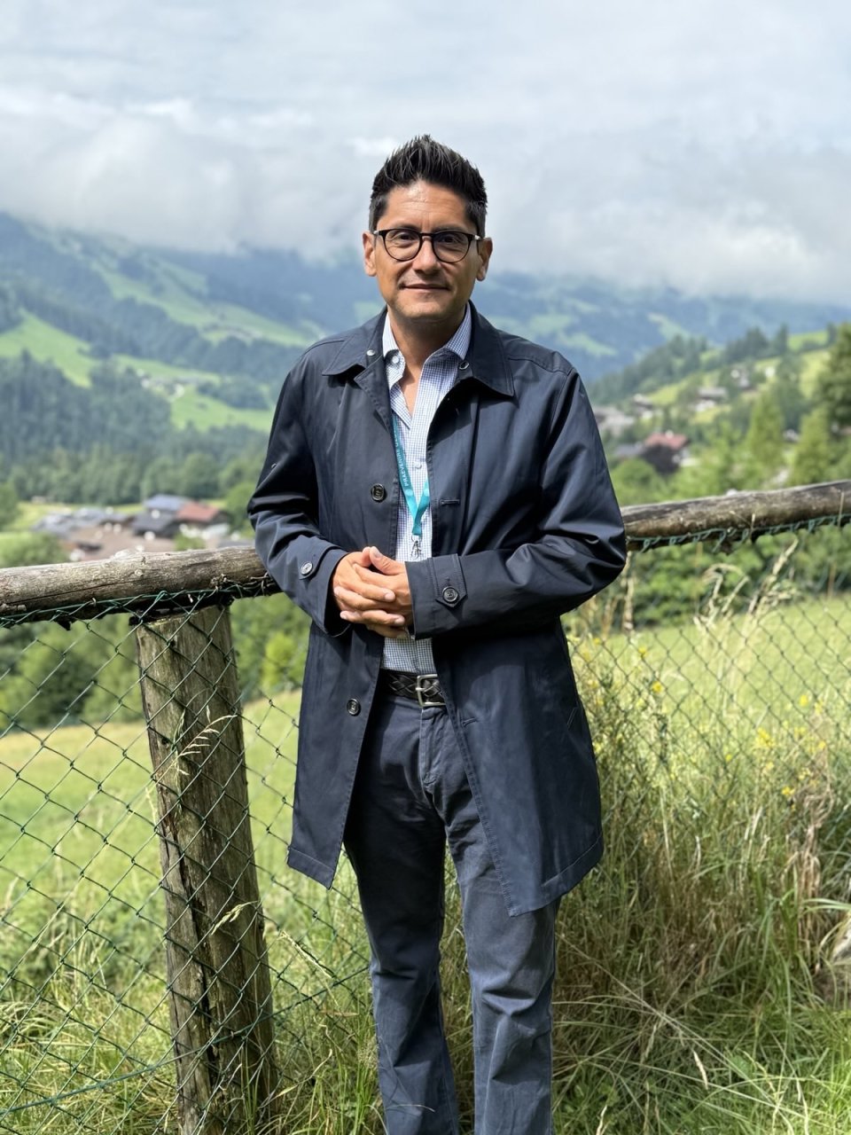 Eduardo Maldonado-González, steht angelehnt an einen Holzzaun und im Hintergrund ist nebelige alpine Landschaft zu sehen. Das Foto wurde im Tiroler Alpbach aufgenommen.