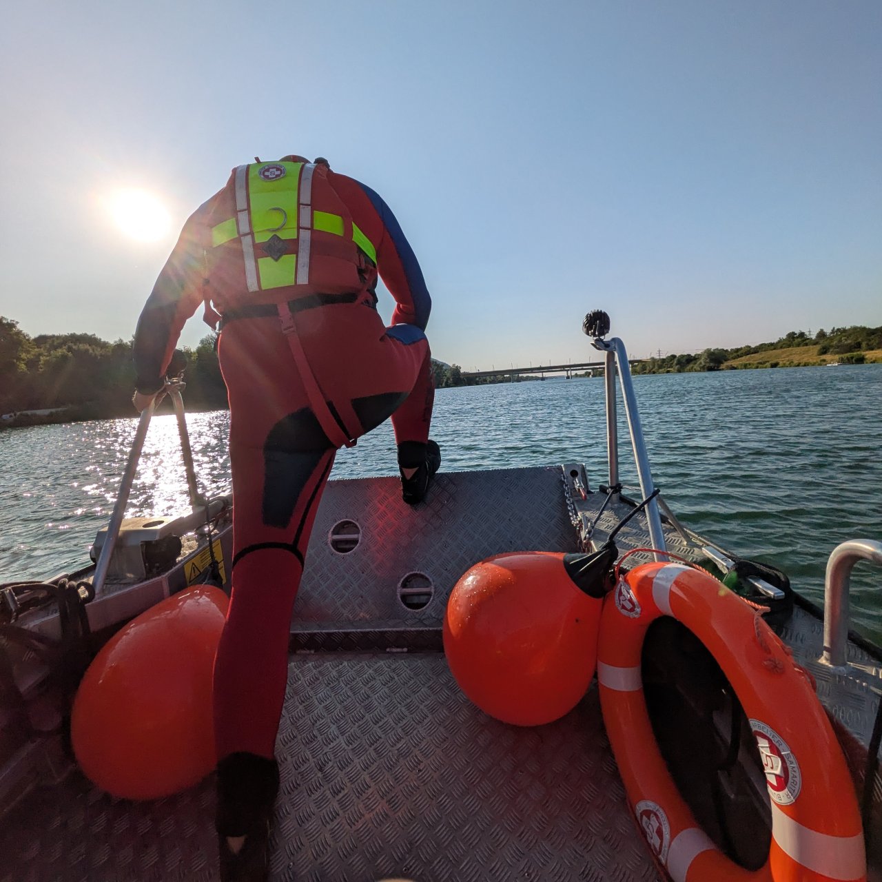 Man sieht eine Einsatzkraft von hinten in der Arbeitskleidung in einem roten Overall steht auf einem Rettungsboot das am Wasser fährt.