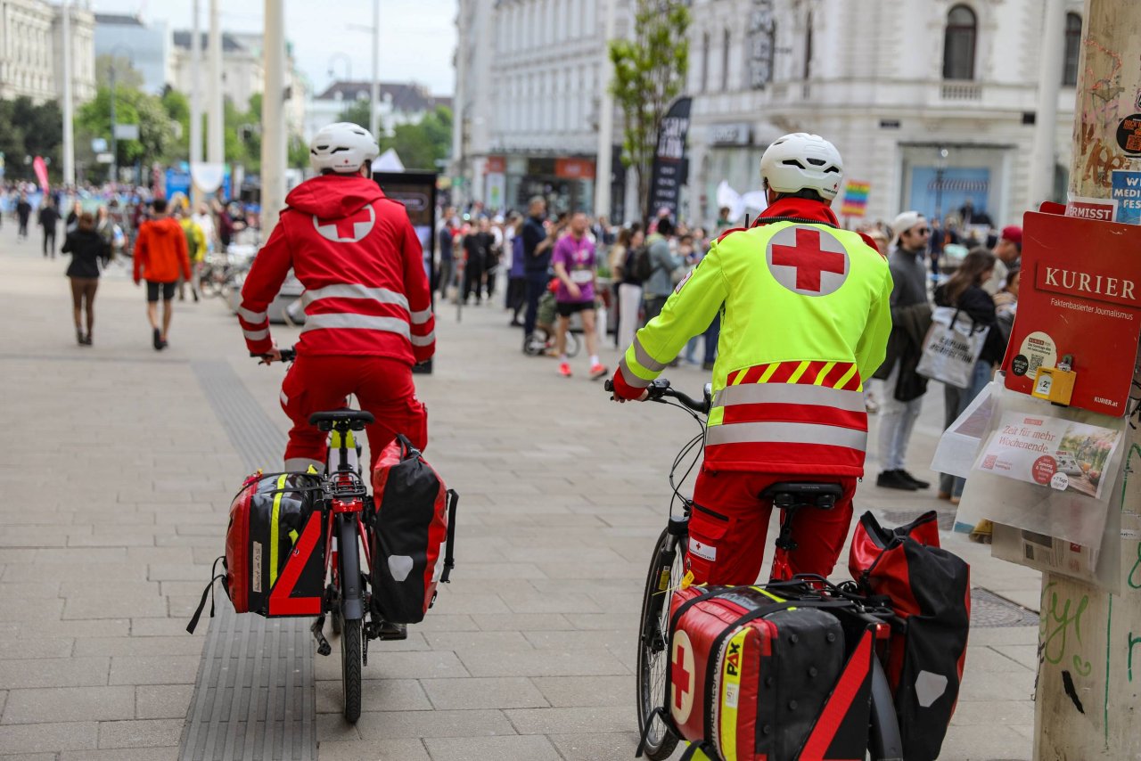 Zwei Rot-Kreuz-Mitarbeiter sieht man von hinten auf E-Bikes sitzen und wie sie gerade die Wiener Mariahilfer Straße hinunter fahren.