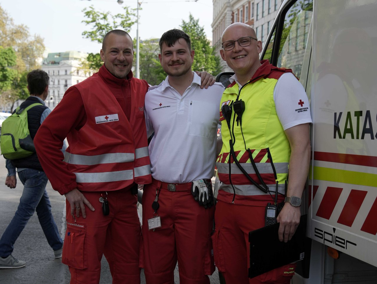 Drei Personen vom Roten Kreuz stehen dicht beieinander bei einem Einsatzfahrzeug.