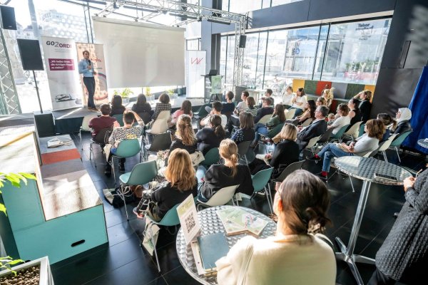 Vortragssaal mit Vortragendem am Podium (stehend) und zahlreichen Personen im Publikum (sitzend).