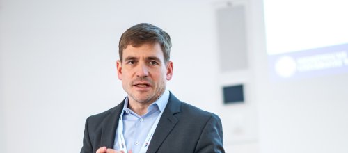 Ein Mann steht bei einem Vortrag und spricht, während er beide Hände vorne zusammenführt beim Sprechen. 