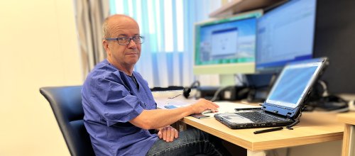 Ein Mann sitzt in einem Sessel vor einem Schreibtisch, auf dem ein Laptop und zwei zusätzliche Bildschirme stehen.