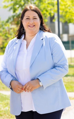 Im Vordergrund ist Frau Kathrin Gaál zu sehen. Sie steht und lächelt in die Kamera. Im Hintergrund sieht man zwar urbane Umgebung, aber mit viel Grün.