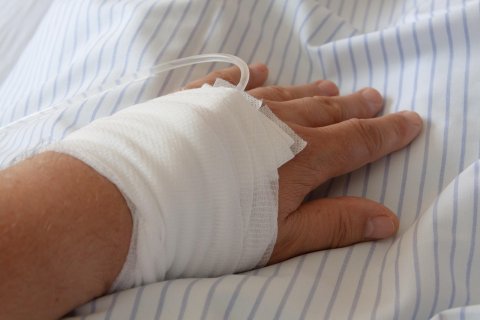Man sieht eine linke Hand, die mit einem Verband versehen ist, aus dem ein Infusionsschlauch herausragt.