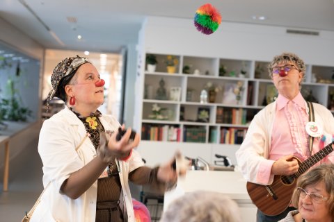 Zwei CliniClowns bei ihrer Performance. Der männliche Clown spielt gerade auf einer kleinen Gitarre/Ukulele und der weibliche Clown jongliert gerade mit einem bunten Ball.