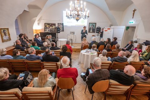 Clubraum mit zahlreichen sitzenden Menschen, die einer Frau mit Mikrofon zuhören