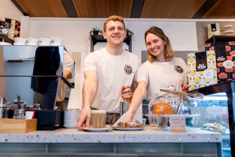 Zwei junge Leute, ein Mann und eine Frau, stehen hinter dem Tresen und haben jeweils die rechte Hand bei einem Gegenstand - Getränk und Teller mit einem Kuchen.