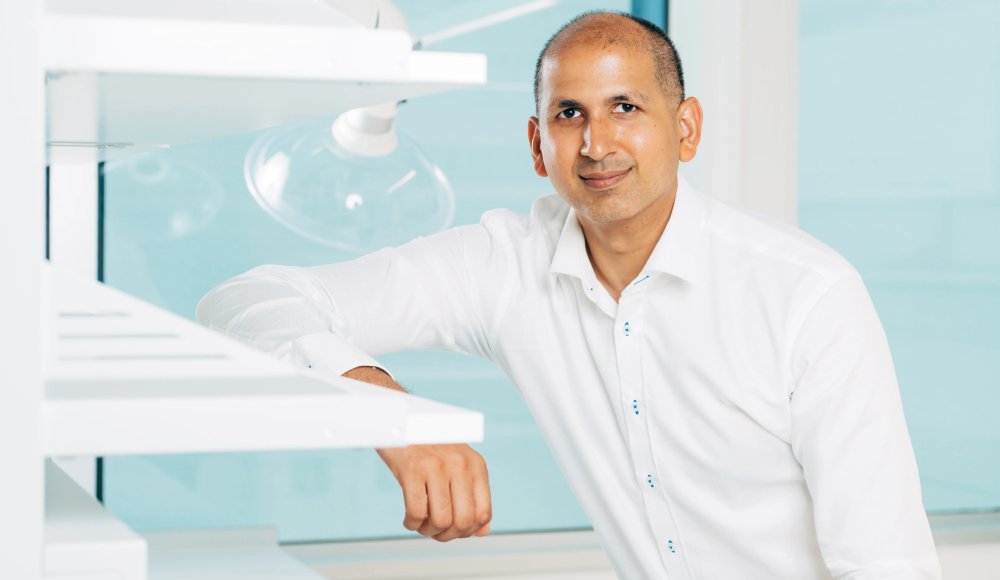 Der Mediziner Wali Malik sitzt in einem Labor, weiß bekleidet und blickt direkt in die Kamera.