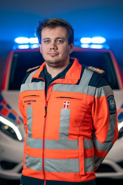 Zu sehen ist der Einsatzleiter der Wiener Berufsrettung, Dominik Kalmar, der eine typische Rettungsjacke trägt - hinter ihm ist ein Rettungswagen zu sehen.