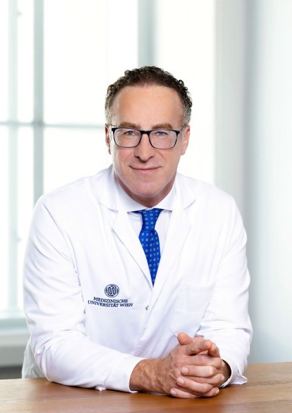 Der Arzt Shahrokh Shariat sitzt an einem Tisch, die Hände vor sich zusammengetan, und hat einen weißen Arztkittel an und trägt eine blaue Krawatte. Er trägt ebenso eine Brille und blickt direkt in die Kamera.