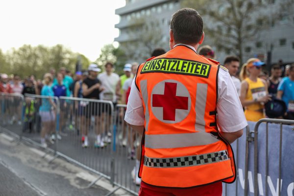 Der Einsatzleiter vom Roten Kreuz steht mit dem Rücken zur Kamera und blickt auf die Läuferinnen und Läufer, die man hinter einer Absperrung auf einer Straße laufen sieht.
