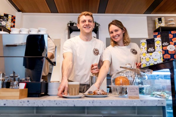 Zwei junge Leute, ein Mann und eine Frau, stehen hinter dem Tresen und haben jeweils die rechte Hand bei einem Gegenstand - Getränk und Teller mit einem Kuchen.