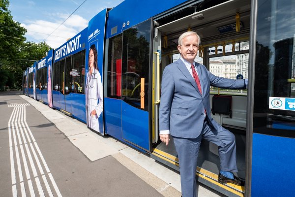 Ein steht in der Tür einer geöffneten Straßenbahn - mit einem Bein noch in der Straßenbahn, mit dem anderen bereits am Trottoir.